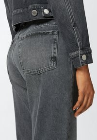 Siva traper jakna s metalnim gumbima, koja ima pripijeni kroj i blago izblijedjeli izgled. Vidljiv je stražnji džep s ukrasnim šavovima.