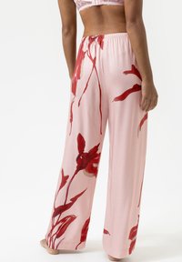 Femme portant un pantalon ample, rose clair avec de grands motifs floraux rouges, debout pieds nus contre un fond blanc uni.