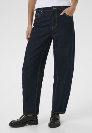 Jeans din denim albastru închis cu o croială lejeră, talie înaltă, cusături contrastante, buzunare frontale și fermoar. Se poartă pantofi negri.