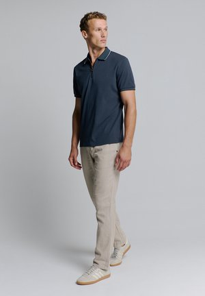 Marineblauwe polo met een ritskraag en witte accenten, gecombineerd met lichtbeige tapered pantalons en witte sneakers met beige details.