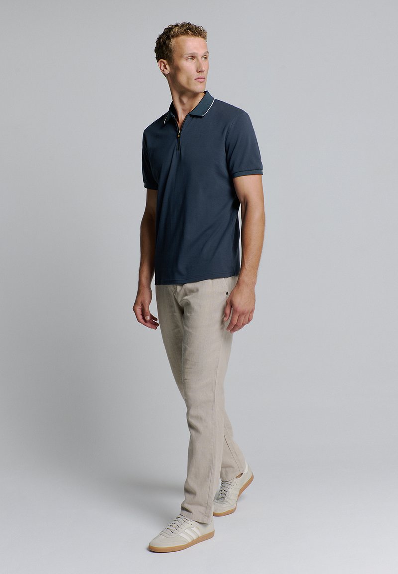 Marineblauwe polo met een ritskraag en witte accenten, gecombineerd met lichtbeige tapered pantalons en witte sneakers met beige details.