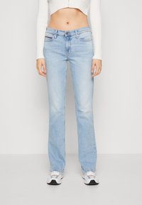 Persona che indossa jeans dritti azzurri chiari, maglietta bianca a maniche lunghe a costine tagliata, e sneakers bianche, in piedi contro uno sfondo semplice.