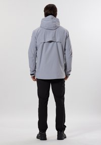 Veste imperméable gris clair avec capuche, poignets ajustables et aération au dos, associée à un pantalon noir et des chaussures noires.