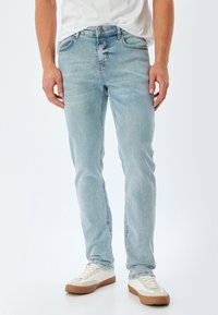 Jeans de color azul claro con corte estándar y una textura descolorida. Presenta cinco bolsillos y un cierre frontal con botón. Llevados con zapatillas blancas.