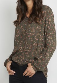 Blouse vert olive avec un motif floral en rose, orange et violet, dotée d'un col en V et de manches longues, fabriquée en tissu léger.