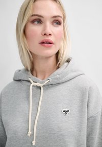 Grå hoodie i mjukts material, med snörning i krämfärg och en liten broderad logga på bröstet.