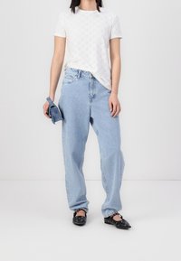 Vit kortärmad skjorta med ögla-mönster, avslappnade blå denimjeans och svarta lägenheter, håller en liten blå väska. En enkel, avslappnad outfit.