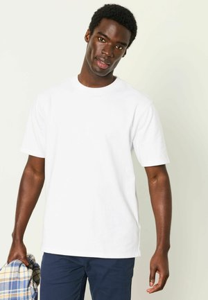 Mand iført en ensfarvet hvid T-shirt og marineblå bukser, der holder en foldet blå og beige ternet skjorte i venstre hånd.