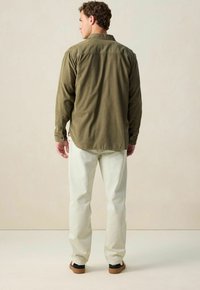 Olijfgroene, knoop-down blouse met een rechte zoom, gecombineerd met lichte broek en donkere schoenen, gezien vanaf de achterkant.