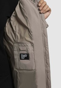 Fliisbeige jakki, millel on sile tekstuur, varustatud lukuga ja nööpidega. Seespool tasku musta sildiga. Must särk on nähtav all.