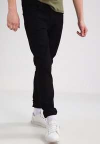 Jeans de denim negro con un corte ajustado, costuras visibles, diseño de cinco bolsillos, usados con una camisa verde claro y zapatillas blancas.