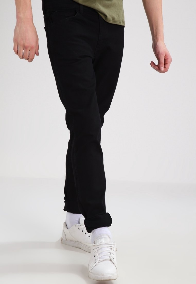 Jeans de denim negro con un corte ajustado, costuras visibles, diseño de cinco bolsillos, usados con una camisa verde claro y zapatillas blancas.