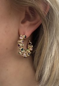 Gouden hoepeloorbelletjes met meerkleurige edelstenen, waaronder roze, groen en paars, met een sierlijk bloemenontwerp en gestructureerde accenten.
