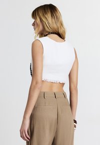 Femme aux cheveux blonds portant un top court sans manches blanc et un pantalon beige taille haute, se tenant devant un fond blanc uni.