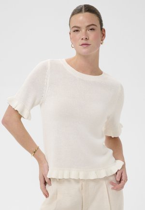 Femme portant un haut en maille blanc à manches courtes avec des bords à volants, un pantalon beige, des boucles d'oreilles créoles dorées et un bracelet en or, posant les mains sur les hanches.