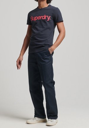 Person iført en marineblå Superdry t-shirt, mørkeblå bukser, hvide sneakers og et sølvarmbånd med den ene hånd i lommen.