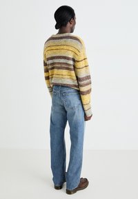 Suéter de punto con rayas amarillas, grises y marrones, combinado con jeans anchos de color azul claro y botas marrones, visto desde atrás.