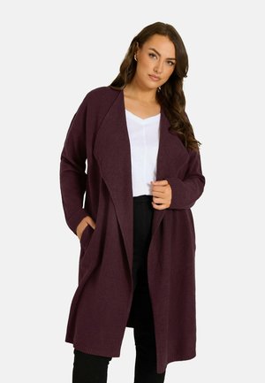 Frau trägt einen langen, dunkelvioletten Cardigan mit offenem Vorderteil über einem weißen Top und schwarzen Hosen, steht vor einem schlichten Hintergrund.