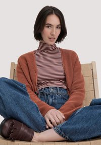 Ein rostfarbener Cardigan über einem gestreiften Rollkragenpullover, kombiniert mit hochgeschnittenen blauen Jeans. Weiche, strukturierte Materialien mit entspannter Passform.