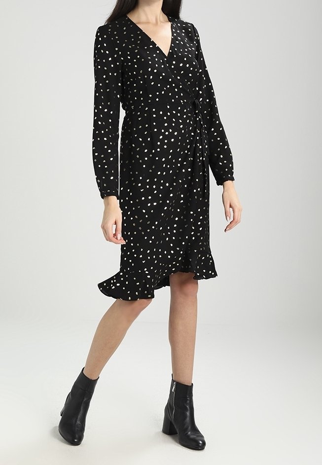Robe noire à pois blancs, à manches longues et avec un ourlet volanté. Portée avec des bottines noires à la cheville, mettant en valeur une silhouette ajustée.