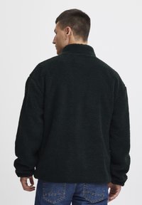 Dark green fleece jacket with a soft, textured finish, high collar, and loose fit. Features dropped shoulders and ribbed cuffs.