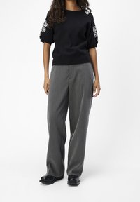 Mujer con suéter negro de manga corta con bordado floral blanco y pantalones anchos grises y sueltos, de pie sobre un fondo blanco.