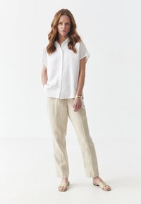 Camicia bianca a maniche corte con bottoni, pantaloni in lino beige e sandali minimalisti beige, che mostrano una vestibilità rilassata e texture morbide.