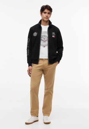 Férfi, aki fekete cipzáras dzsekit visel fehér felirattal, fehér grafikás pólót, bézs nadrágot és fehér sportcipőt, miközben az egyik kezét a zsebében tartja.