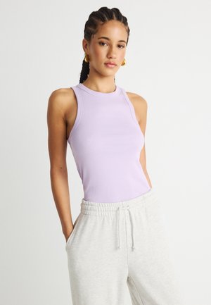 Ung kvinde med fletninger, der bærer guldringeøreringe, en lavendelfarvet ærmeløs top og lysegrå joggingbukser med hænderne i lommerne.