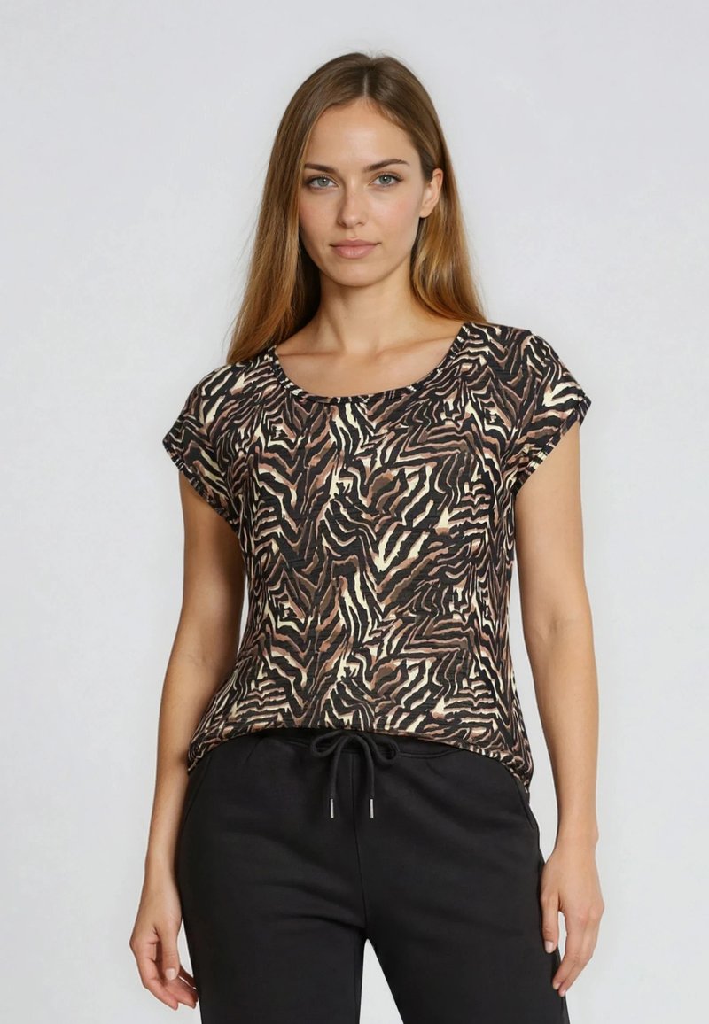 Jeune femme aux longs cheveux portant un haut à manches courtes imprimé animalier marron, beige et noir, et un pantalon noir à cordon, debout devant un fond blanc.