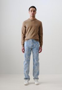 Suéter marrón de manga larga con un pequeño logo, combinado con jeans de camuflaje azul claro y zapatillas blancas, de pie contra un fondo neutro.