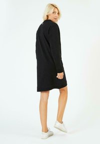 Robe-sweat noire avec des manches longues, un col rond et une coupe au genou. Texture douce, couture minimale et un petit détail accentué à l'arrière.