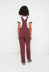 Salopette bordeaux avec des bretelles réglables, jambes larges et deux poches arrière, portée sur un t-shirt blanc à contour noir. Tissu texturé.