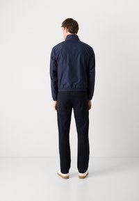 Navy jacket with elastic cuffs and hem, made of water-resistant material, paired with dark trousers and white shoes, viewed from behind.