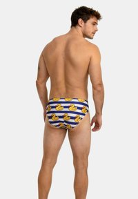 Joven con la espalda al aire que lleva puesta una malla de baño a rayas azul y blanca con un estampado de patitos de goma amarillos, de pie contra un fondo liso.