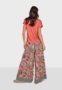 Frau mit dem Rücken zur Kamera, trägt ein korallenfarbenes Kurzarmoberteil und weite Hosen mit roten, beigen, schwarzen Blumen- und Paisley-Mustern auf weißem Hintergrund.