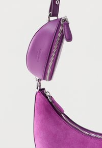 Violetti mokkanahkainen käsilaukku, jossa on sileä nahkainen yksityiskohta, kaareva muoto, hopeiset vetoketjut ja irrotettava pieni säilytyspussi.