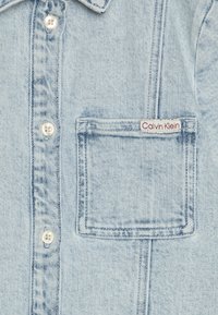Gaiši zils denima krekls ar apkakli, priekšējiem pogām un krūšu kabatu ar izšūtu Calvin Klein logo. Audumam ir teksturēta apdare.