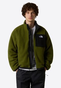 Chaqueta de forro polar verde con cremallera frontal, bolsillo negro y el logo de The North Face. Presenta un corte holgado y una textura suave, con puños en las muñecas.