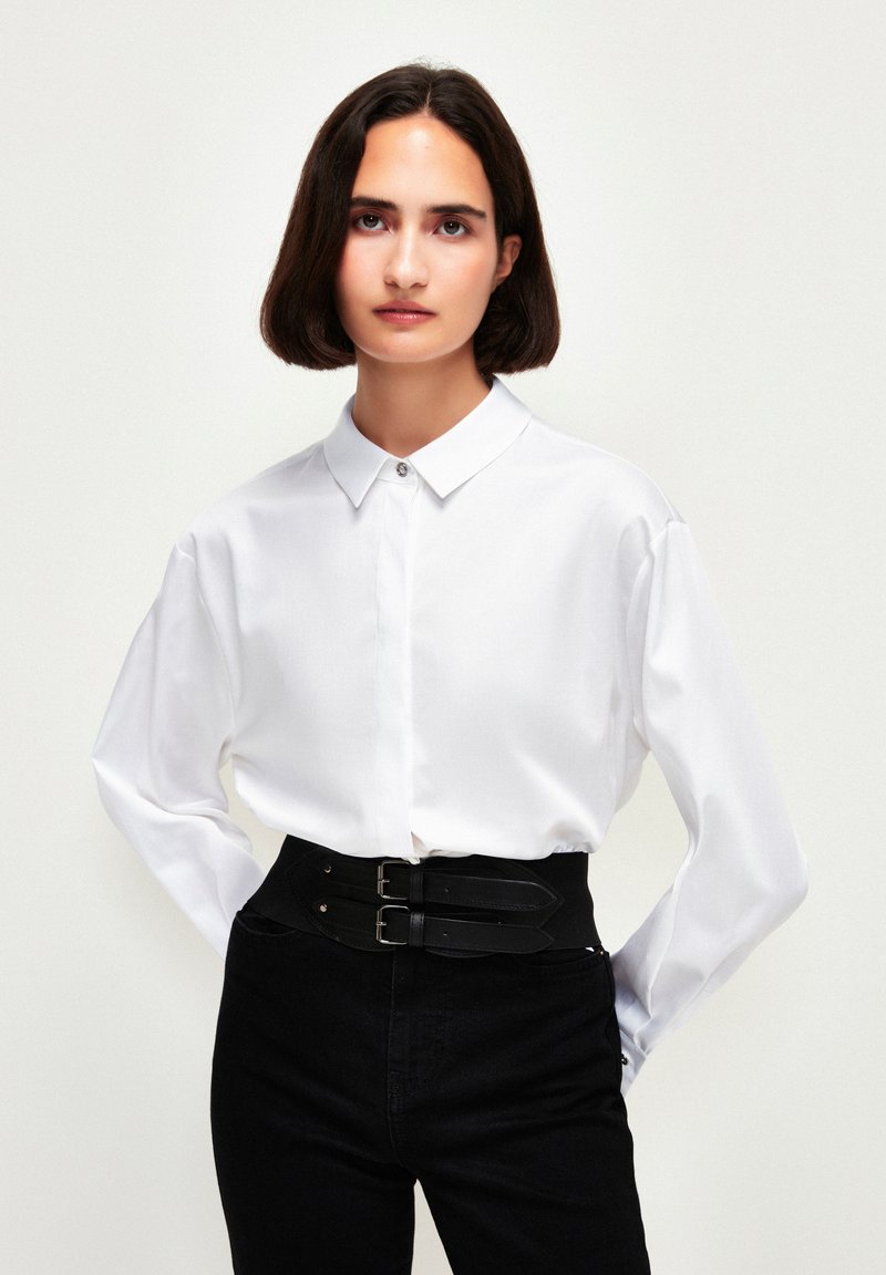 Camicia bianca a maniche lunghe con colletto a punta e placket con bottoni, abbinata a pantaloni neri a vita alta con cintura e doppi fibbie.