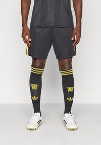 Μαύρο αθλητικό σορτς με κίτρινες πλευρικές λεπτομέρειες, συνδυασμένο με μαύρες κάλτσες μέχρι το γόνατο που έχουν κίτρινες σχέσεις και λευκά ποδοσφαιρικά παπούτσια.