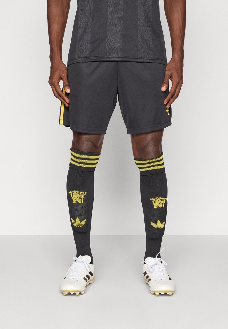 Μαύρο αθλητικό σορτς με κίτρινες πλευρικές λεπτομέρειες, συνδυασμένο με μαύρες κάλτσες μέχρι το γόνατο που έχουν κίτρινες σχέσεις και λευκά ποδοσφαιρικά παπούτσια.