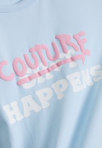 Gaiši zila kokvilnas T-krekls ar teksturētu rozā un baltu printu, kas lasāms "COUTURE HAPPENS" ar rozā pārsvītrojumu.