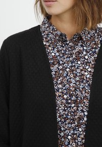 Cardigan nero testurizzato indossato sopra una camicia a fantasia floreale nei toni del blu, bianco e ruggine. Primo piano che evidenzia i dettagli del tessuto.