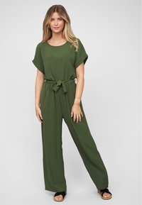 Woman wearing an olive green short-sleeve jumpsuit with a waist tie and black sandals, standing against a plain white background.
