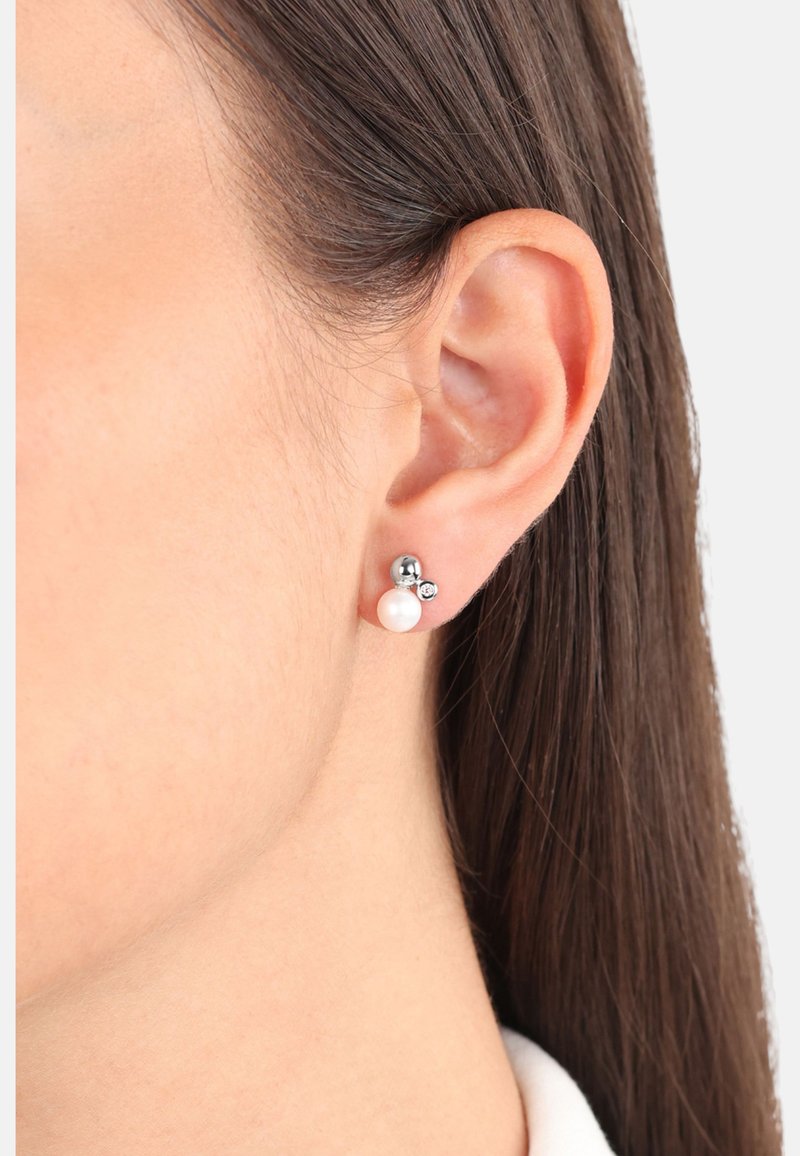 Oreja de mujer con cabello castaño oscuro, lleva pendientes de arete de plata con una perla y una pequeña gema clara.