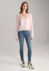 Lichtroze cardigan met een V-hals, voorzien van vijf gouden knopen aan de voorkant. Draagt over een witte tanktop en gecombineerd met blauwe jeans.