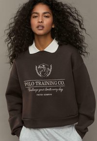 Felpa marrone con colletto bianco, con ricamo della scritta "Polo Training Co." e un logo sul davanti. Tessuto morbido e strutturato.