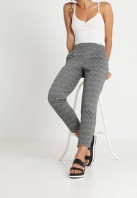 Šedé kalhoty s vzorem a úzkým střihem s elastickým pasem, kombinované s bílým tílkem a černými sandály na bílém pozadí.