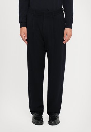 Pantalons plissés noirs avec une texture lisse, coupe légèrement décontractée et passants de ceinture, associés à des chaussures noires brillantes.